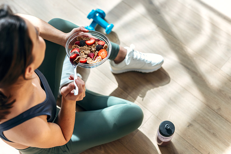 woman in exercise cloths sitting on the floor eating cereal and berries
